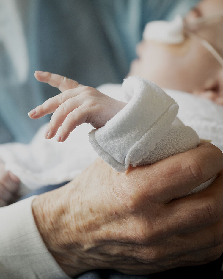 Hand einer älteren Person stützt den Arm eines kleinen Babys, das beatmet wird